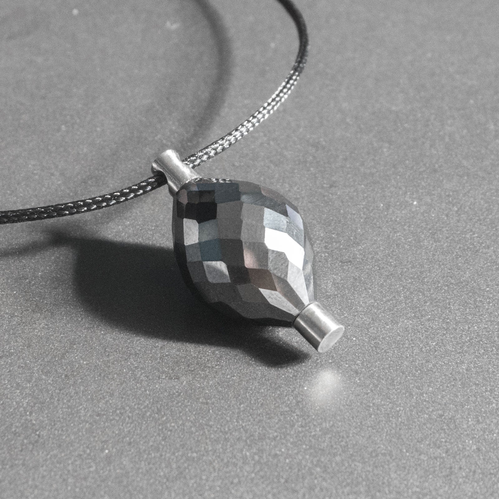 A giant natural black diamond, weighing close to 35cts, is enclosed by 2 cylindrical titanium mounts, making a seamless and continuous shape. Despite the many facets, the pendant looks very subdued.