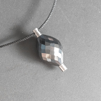 A giant natural black diamond, weighing close to 35cts, is enclosed by 2 cylindrical titanium mounts, making a seamless and continuous shape. Despite the many facets, the pendant looks very subdued.